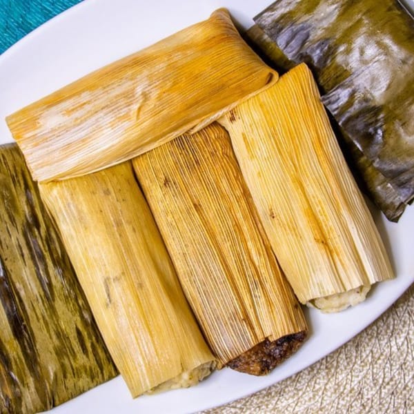 A white plate holds several tamales, some wrapped in corn husks and others in banana leaves.