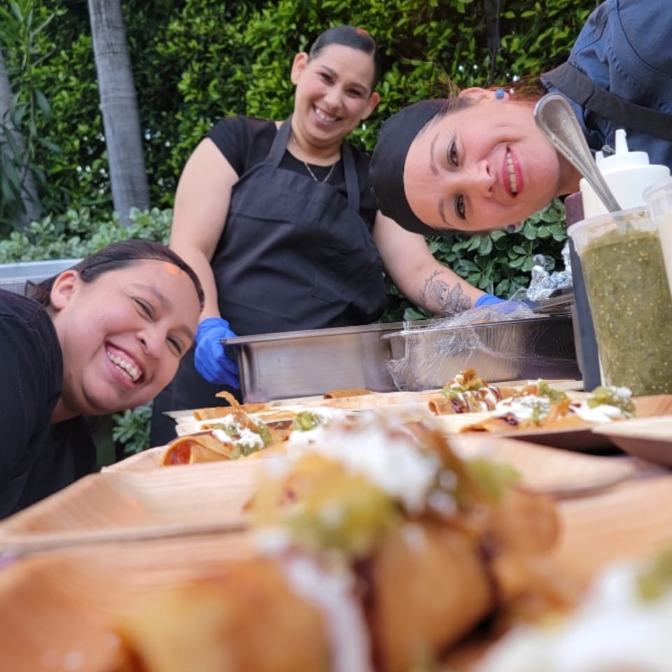Three smiling Loteria Grill chefs behind a table of Mexican taquitos with salsa and crema.