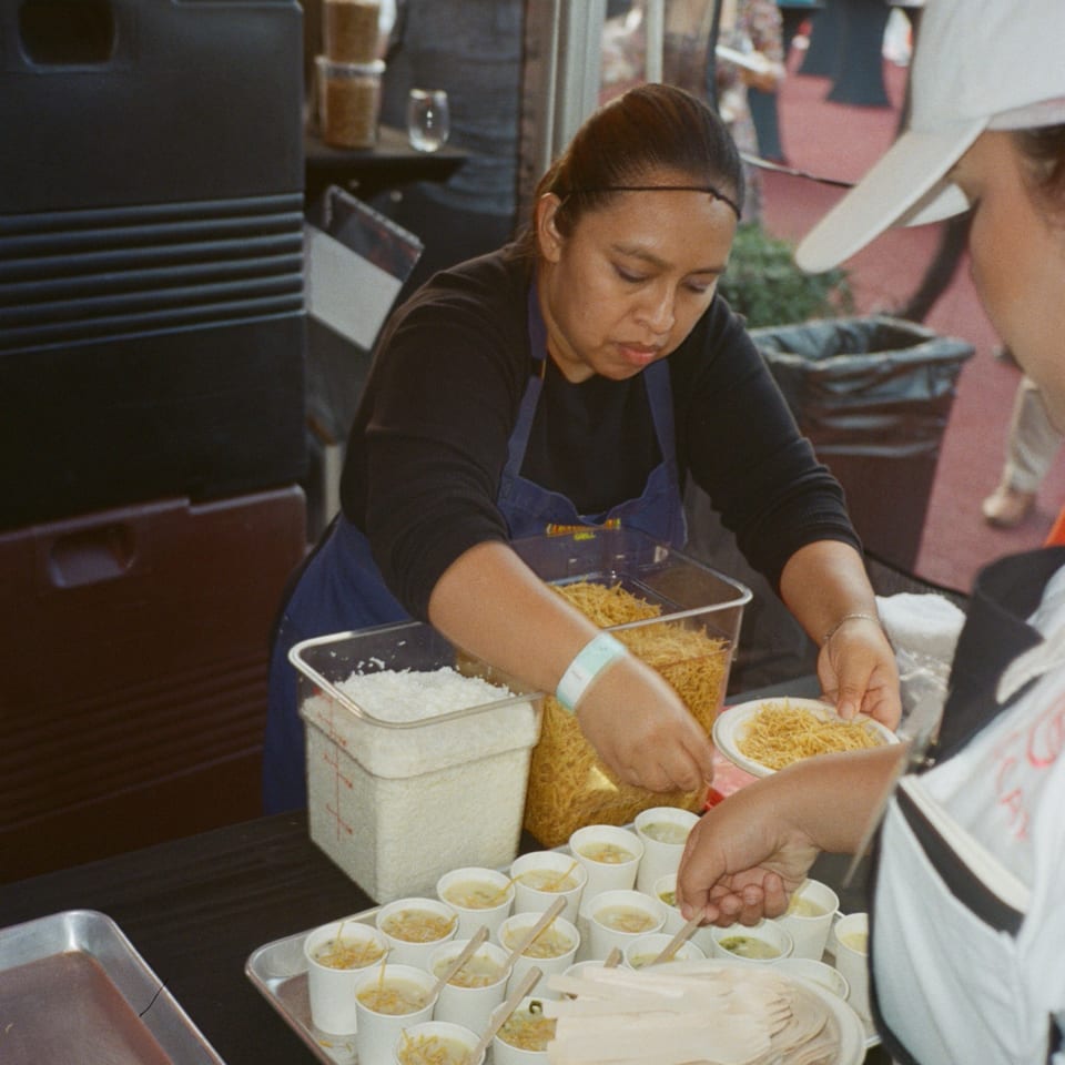 A Loteria Grill employee serving small cups of Mexican street corn with crispy noodles to a customer.