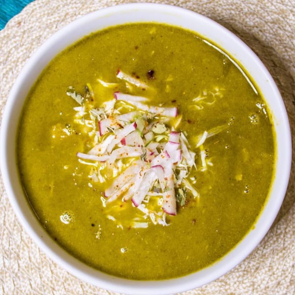 A bowl of green pozole soup, garnished with shredded radishes and cabbage, on a woven placemat.
