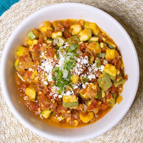 Close-up of a white bowl filled with zucchini, tomatoes, onions, and cheese on a woven placemat.