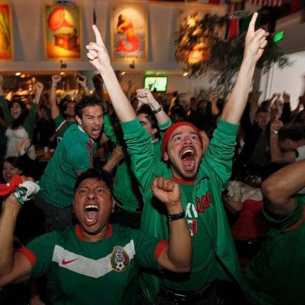 Excited crowd in green jerseys cheering with arms raised at Loteria Grill, celebrating a Mexican sports victory.