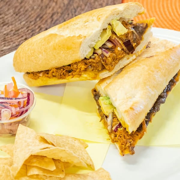 A Mexican torta sandwich, cut in half, with chips and pickled onions on a white plate.