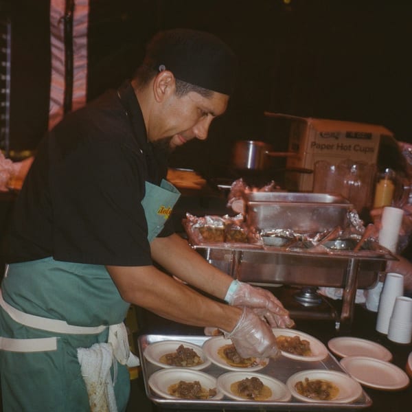 A chef in a black shirt and green apron prepares small Mexican tacos on a tray.