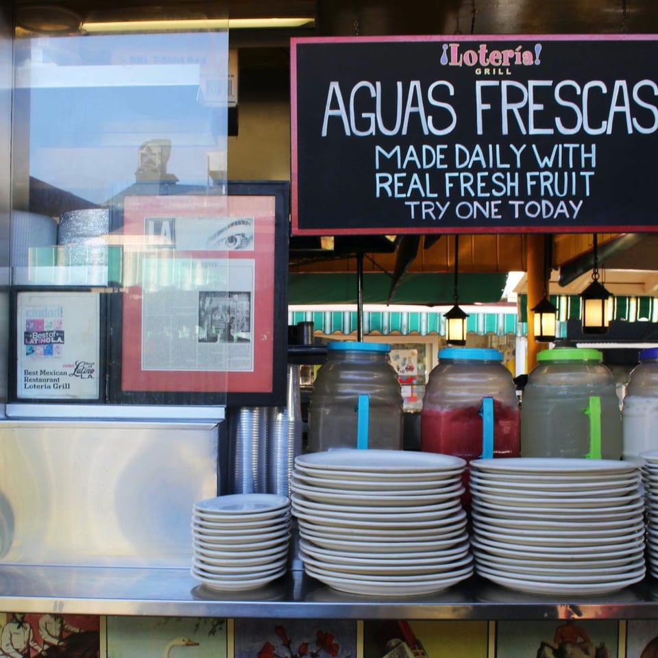 Loteria Grill's Aguas Frescas sign, with large drink dispensers and stacks of plates below.